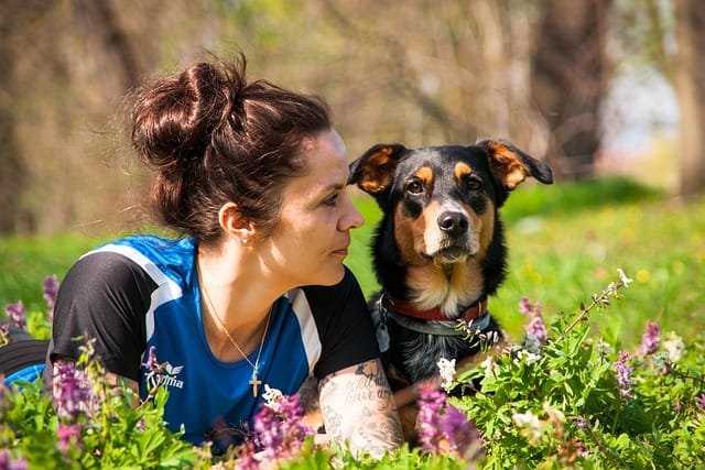 femme regardant de côté et son chien couchés dans l'herbe fleurie face à l'objectif arrière plan flou d'arbres
